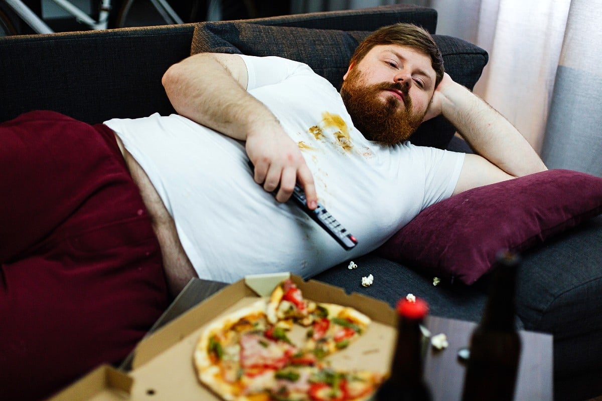 Gordo perezoso comiendo pizza tumbado en el sofá viendo la tele.